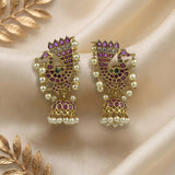 A pair of gold-plated Indian jhumka earrings decorated with purple and green stones and white faux pearls, displayed on a beige satin fabric with gold leaf accents.