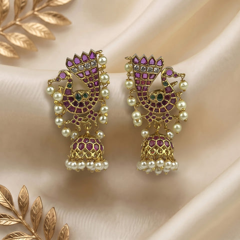 A pair of gold-plated Indian jhumka earrings decorated with purple and green stones and white faux pearls, displayed on a beige satin fabric with gold leaf accents.