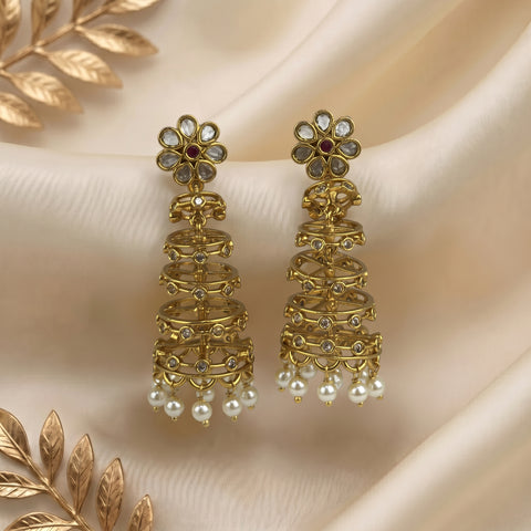 A pair of ornate gold-toned jhumka earrings with floral studs, clear stone accents, and dangling pearl-like beads, displayed on draped cream fabric with decorative gold leaf elements.