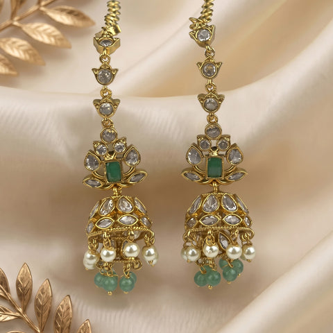 A pair of gold-toned traditional Indian jhumka earrings decorated with clear kundan stones, green rectangular stones, and white pearl-like beads, displayed on draped cream satin fabric with decorative gold leaves.