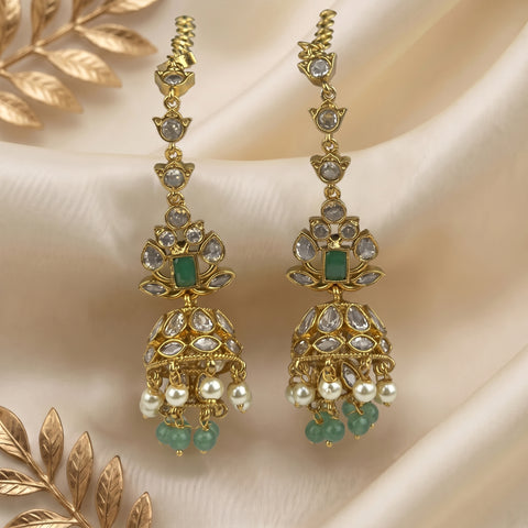 A pair of ornate gold-toned dangle earrings with clear kundan stones, green rectangular inlays, and hanging pearl-like beads, displayed on draped cream satin fabric with decorative gold leaves.