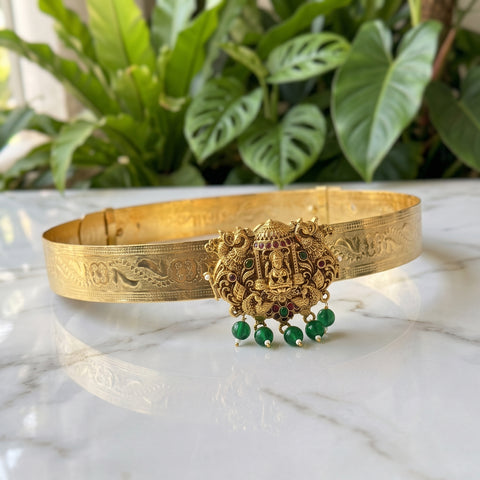An ornate gold-toned traditional collar or waist chain with intricate engraved patterns and a central deity motif, accented with small green beads, displayed on a white marble surface with a blurred background of green plants.