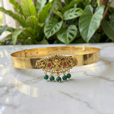 Gold-toned traditional Indian choker necklace with a central ornate filigree piece featuring red stones and dangling green bead tassels, displayed on a white marble surface with green plants in the background.