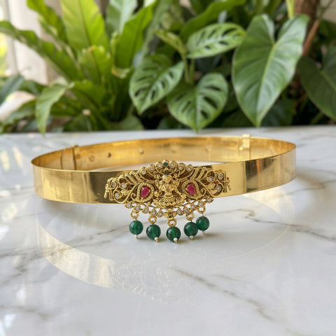 Gold-toned traditional Indian choker necklace with a central ornate filigree piece featuring red stones and dangling green bead tassels, displayed on a white marble surface with green plants in the background.