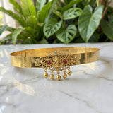 A gold-toned traditional Indian choker necklace with intricate filigree work, red gemstone accents, and dangling gold bells, displayed on a white marble surface with green plants in the background.