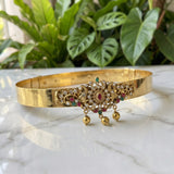 A gold-plated traditional Indian choker bangle with intricate rhinestone and gemstone embellishments, resting on a white marble surface with a blurred background of green houseplants.