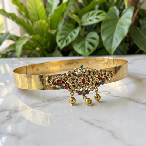A gold-plated traditional Indian choker bangle with intricate rhinestone and gemstone embellishments, resting on a white marble surface with a blurred background of green houseplants.