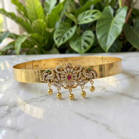 A gold-plated traditional Indian choker necklace with a highly detailed central filigree motif, set with small green and red stones and three dangling gold bells, displayed on a white marble surface with a blurred green plant background.