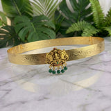 An ornate gold-toned traditional headband or tiara with engraved patterns and a central deity motif, accented with small green gemstones and dangling green bead drops, displayed on a marble surface with green foliage in the background.