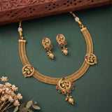 Gold-toned traditional necklace and matching earrings with red and green stone accents, arranged on a dark green background with small dried flowers and a carved wooden prop.