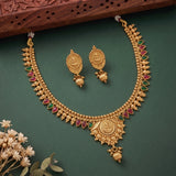 Gold-toned traditional Indian jewelry set featuring a necklace and matching earrings with red and green stone accents, arranged on a dark green background with dried flowers and a carved wooden prop.