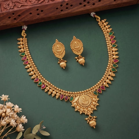 Gold-toned traditional Indian jewelry set featuring a necklace and matching earrings with red and green stone accents, arranged on a dark green background with dried flowers and a carved wooden prop.