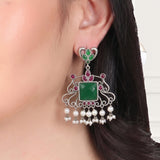 A close-up of a woman's ear wearing a large silver-toned dangle earring decorated with a green stone, pink stones, and dangling white pearl beads.