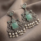 A pair of ornate silver-toned dangle earrings with light green faceted stones and hanging pearl-like beads, displayed on draped taupe fabric.