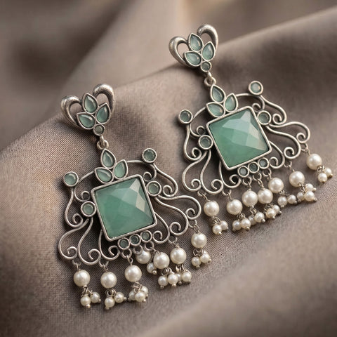 A pair of ornate silver-toned dangle earrings with light green faceted stones and hanging pearl-like beads, displayed on draped taupe fabric.