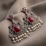 A pair of ornate silver-toned dangle earrings with deep red faceted stones and dangling white pearl-like beads, displayed on draped taupe fabric.