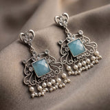 A pair of ornate silver-toned dangle earrings with faceted light blue center stones and dangling pearl-like beads, displayed on draped taupe fabric.