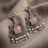 A pair of ornate silver-toned dangle earrings with pink square stones and dangling pearl-like beads, displayed on draped taupe fabric.