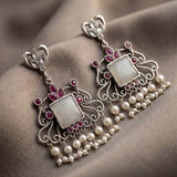 A pair of ornate silver-toned dangle earrings with white pearl-like beads, pink gemstone accents, and square white inlays, displayed on draped taupe fabric.