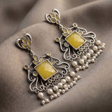 A pair of ornate silver-toned Indian-style earrings with yellow gemstone centers and dangling pearl beads, displayed on a textured taupe fabric background.