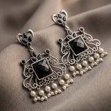 A pair of ornate silver-toned jhumka earrings with black faceted stones and dangling pearl-like beads, displayed on draped taupe fabric.