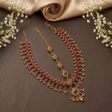 Gold-toned traditional Indian jewelry set including a necklace, earrings, and bangles, decorated with red and green stones, arranged on a dark brown surface with white baby's breath flowers and a beige satin cloth.