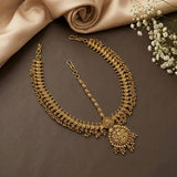 An ornate gold-toned traditional necklace with a large pendant and matching chain laid flat on a dark brown surface, styled with beige satin fabric and small white flowers.