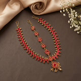 Gold-toned traditional Indian jewelry set with red stones, including a necklace, earrings, and a maang tikka, arranged on a dark brown surface with beige satin fabric and small white flowers.