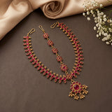 Gold-toned traditional Indian jewelry set featuring a necklace, matching earrings, and a maang tikka, embellished with deep red stones, arranged on a dark brown surface with beige satin fabric and white baby's breath flowers.