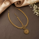 An ornate gold-toned traditional necklace set with a matching chain and a large circular pendant, arranged on a dark brown surface with beige satin fabric and small white flowers as styling props.
