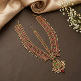 An ornate gold-toned Indian bridal necklace (choker) with matching belly band, decorated with red and green enamel, white pearls, and a red bead pendant, styled on a dark brown surface with beige fabric and baby's breath flowers.