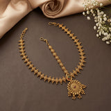 Gold-toned traditional Indian jewelry set featuring a necklace with a large circular sunburst pendant, a matching chain, and a small matching charm, arranged on a dark brown surface with beige satin fabric and white baby's breath flowers.