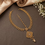 An ornate gold-toned traditional necklace with a matching chain and pendant, styled on a dark brown surface with beige satin fabric and small white flowers.