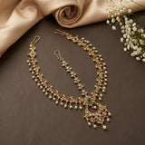 Gold-toned traditional Indian jewelry set featuring a necklace, matching earrings, and a maang tikka, embellished with small white pearls and a central green stone, arranged on a dark brown surface with beige satin fabric and white baby's breath flowers.