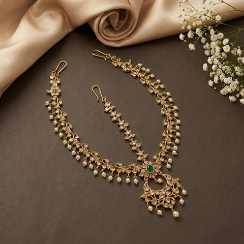Gold-toned traditional Indian jewelry set featuring a necklace, matching earrings, and a maang tikka, embellished with small white pearls and a central green stone, arranged on a dark brown surface with beige satin fabric and white baby's breath flowers.