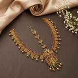 An ornate gold-toned traditional necklace set with red and green stone accents, displayed on a dark brown surface with beige satin fabric and small white flowers.