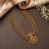 An ornate gold-toned traditional necklace set with a matching pendant and earrings, decorated with red and green stone accents, arranged on a dark brown surface with beige fabric and small white flowers.