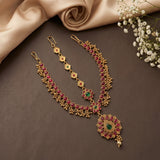 Gold-toned traditional Indian jewelry set including a necklace, matching earrings, and a maang tikka, decorated with red and green stones, arranged on a dark brown surface with beige satin fabric and white baby's breath flowers.