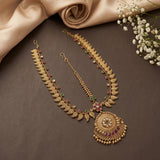 Gold-toned traditional Indian jewelry set featuring a necklace with a large ornate pendant, a matching chain, and a maang tikka, decorated with small green and pink gemstones, displayed on a dark brown surface with beige satin fabric and white baby's breath flowers.