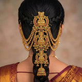 A woman viewed from the back wearing an ornate gold hair ornament with red and green stone accents, styled over a long black braid.