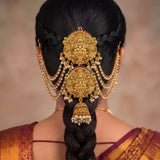 A close-up rear view of a woman wearing an ornate gold traditional Indian hair ornament (jhumka) with hanging pearl strands and a dangling bell, styled with a dark braided hair and a gold and purple sari.