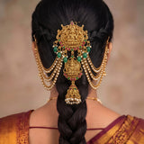 A close-up rear view of a woman wearing an ornate gold-toned traditional Indian hair ornament (jhumka) with hanging pearl strands, green and red gemstone accents, and a large central deity motif. Her hair is styled in a long braid, and she wears a gold and maroon sari against a soft beige background.