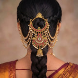 A close-up back view of a woman wearing an ornate gold hairpiece with red and green stone embellishments and dangling pearl-like chains, styled with a long dark braid.
