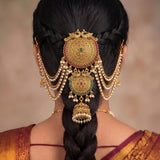 A woman viewed from behind wearing an ornate traditional Indian gold hair ornament (manglik) with intricate filigree, hanging pearl strands, and small red and green stone accents, styled in a deep side braid.