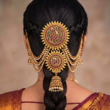 A close-up back view of a woman wearing an elaborate traditional gold hair ornament with red and green stone detailing, displayed on her dark hair.