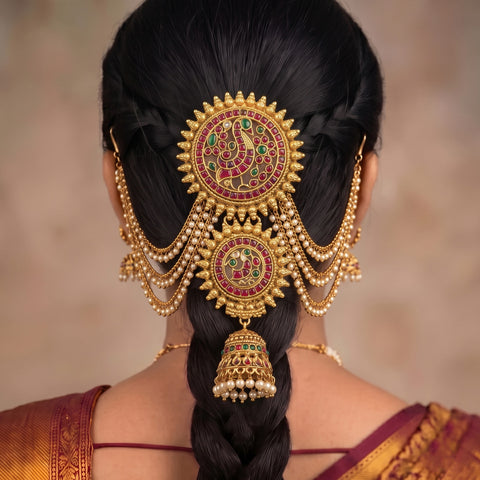 A close-up back view of a woman wearing an elaborate traditional gold hair ornament with red and green stone detailing, displayed on her dark hair.