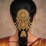 A close-up rear view of a woman wearing an ornate traditional Indian gold hair ornament (jhumka) with red and green stone accents and dangling pearl beads on her dark hair.
