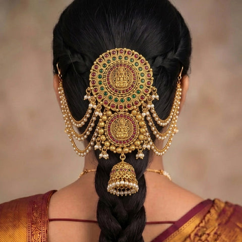A close-up rear view of a woman wearing an ornate traditional Indian gold hair ornament (jhumka) with red and green stone accents and dangling pearl beads on her dark hair.
