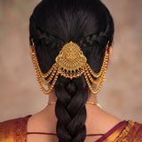 A close-up rear view of a woman wearing an ornate gold traditional hair ornament with dangling beaded chains and a central decorative piece, styled with a long dark braid.