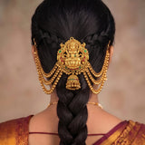A close-up back view of a woman wearing an ornate gold traditional Indian hair ornament (jhumka) with hanging beaded chains and a central deity motif, styled with a long dark braid.